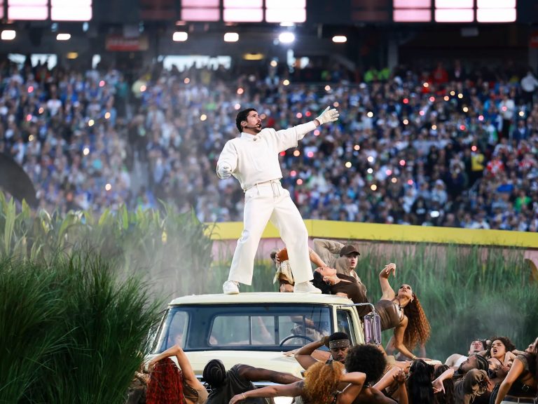 Bad Bunny dressed in white standing on the roof of a vintage vehicle during a stadium performance, surrounded by dancers and a large illuminated crowd.