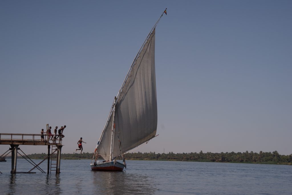 Niños egipcios saltando al río Nilo junto a una faluca tradicional en el Alto Egipto, símbolo de infancia, comunidad y futuro.