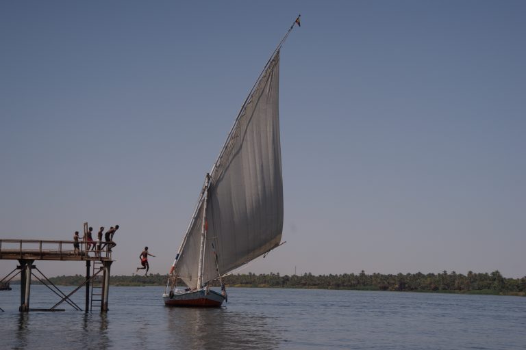 Niños egipcios saltando al río Nilo junto a una faluca tradicional en el Alto Egipto, símbolo de infancia, comunidad y futuro.