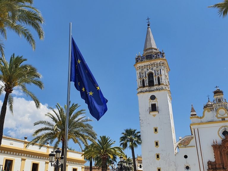 Bandera de la Unión Europea en una plaza de La Palma, con palmeras y una torre de iglesia blanca bajo el cielo azul.