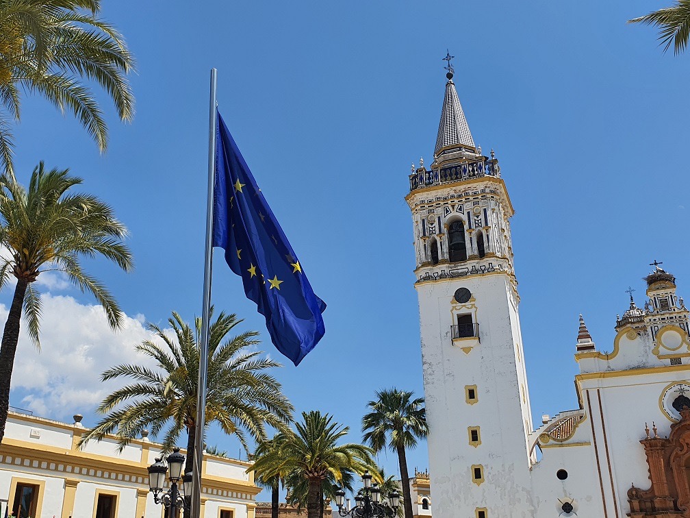 Bandera de la Unión Europea en una plaza de La Palma, con palmeras y una torre de iglesia blanca bajo el cielo azul.